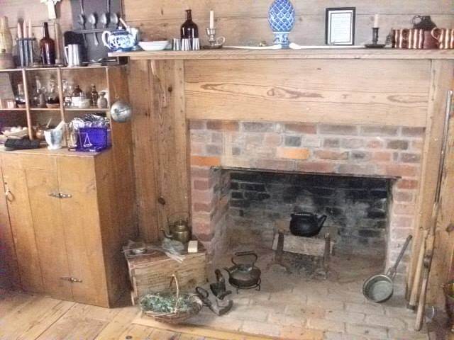A rustic room with a brick fireplace. Various antique items, such as kettles, candle holders, a lantern, and dishware, are displayed on a wooden mantle and shelves. The floor around the fireplace also has vintage objects, including a basket and a pair of old shoes.