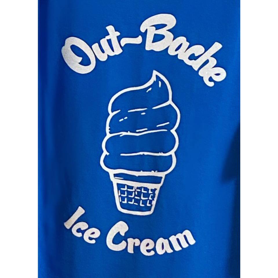 A blue shirt with a white graphic design featuring a soft-serve ice cream cone in the center. Above the ice cream, it says 