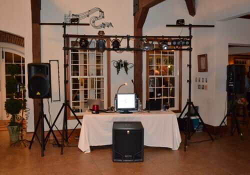 A DJ setup at an indoor event features a table with a laptop, microphones, and speakers. Lighting equipment is mounted on a frame above the table, and additional speakers are on stands beside the table. The room has large windows and wood beams. Image