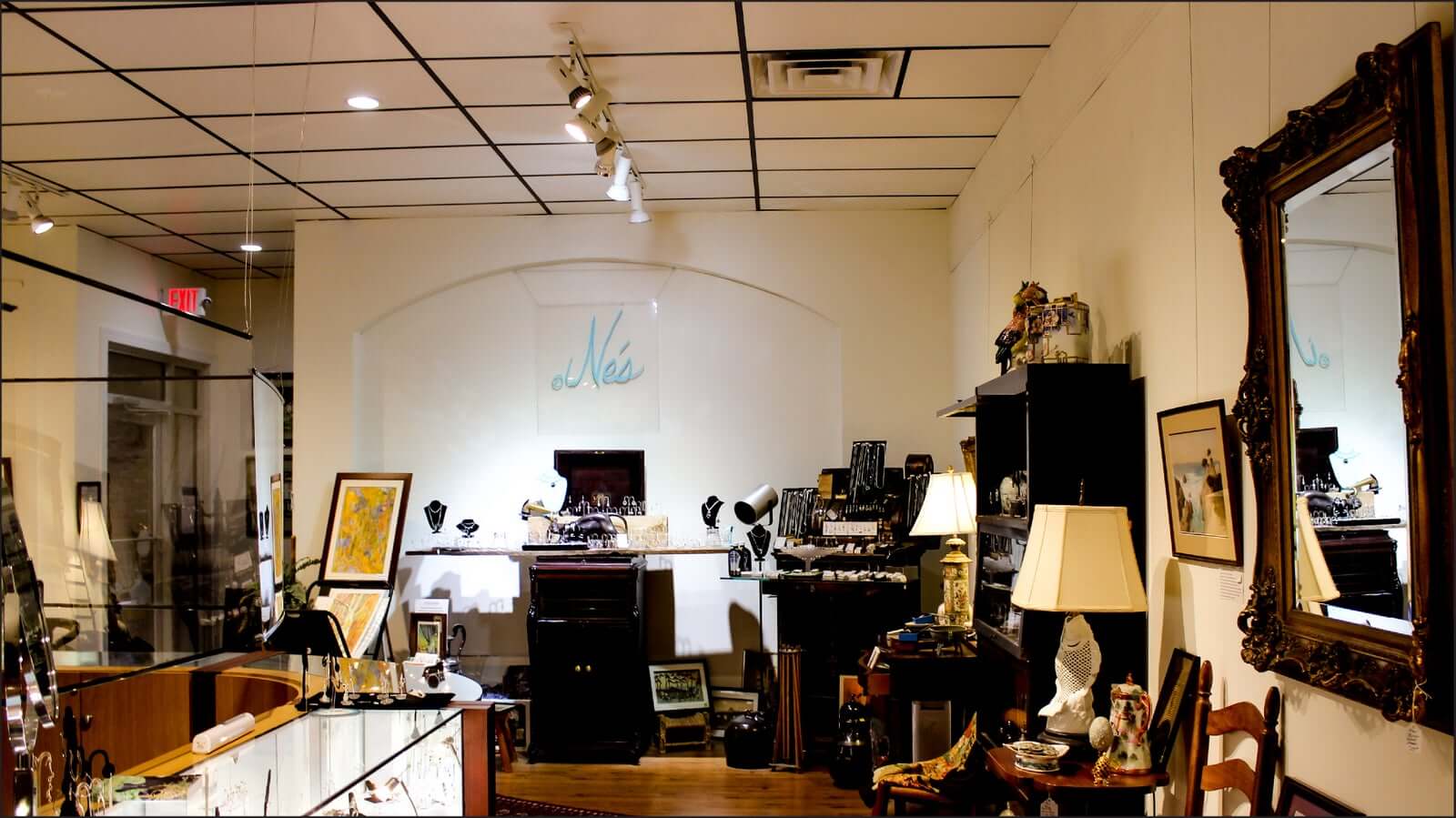 A small, well-lit antique shop with wall decor, glass display cases, various framed pictures, lamps, and vintage items. A large mirror and typewriters can be seen, while a sign