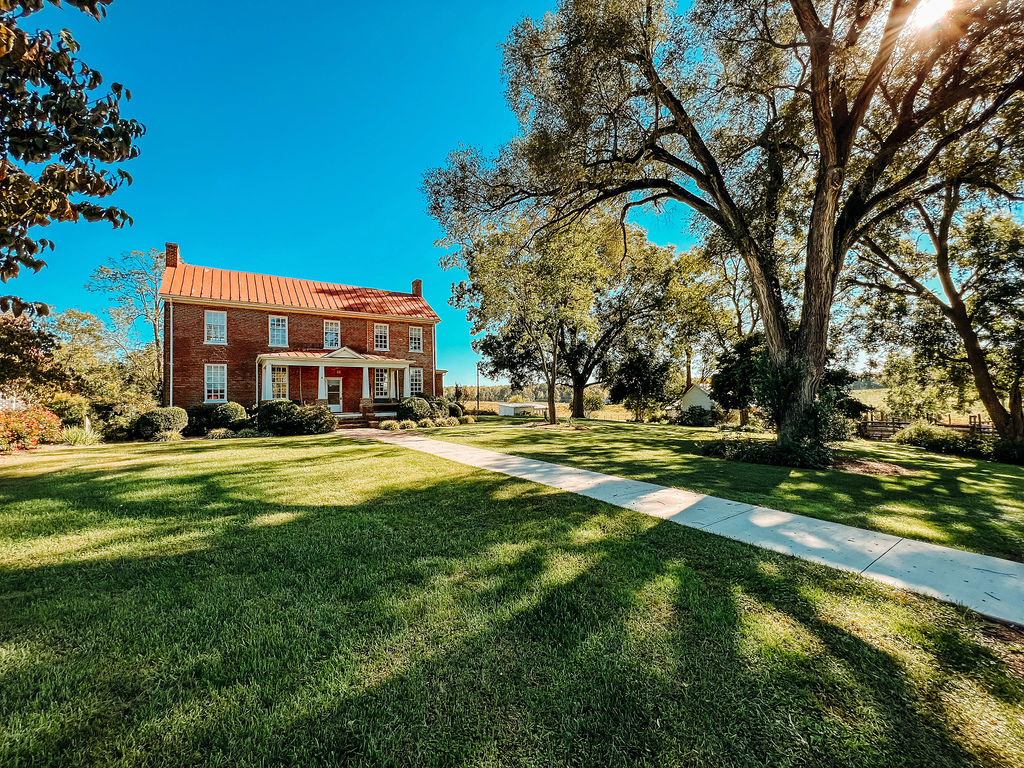 A two-story brick house with a red roof and white-trimmed windows and doors stands at the end of a long, straight sidewalk. The house is surrounded by a well-maintained lawn and large trees, casting long shadows across the green grass. The sky is clear and blue.