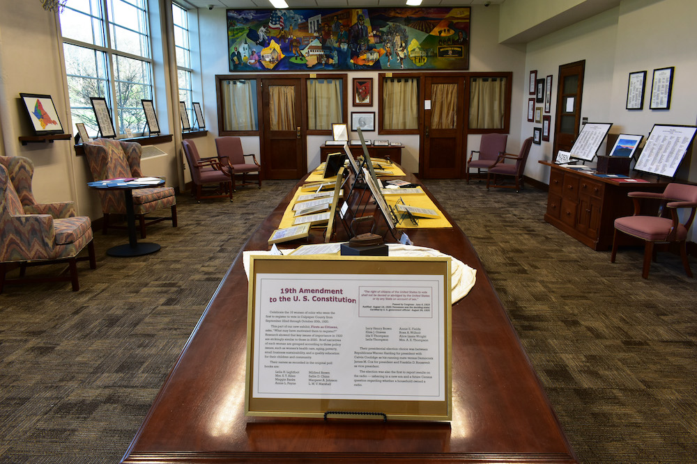 A room with a large wooden table in the center displaying various historical documents, including an informational plaque about the 19th Amendment to the U.S. Constitution. The room has chairs, large windows, framed artwork, and shelves along the walls.