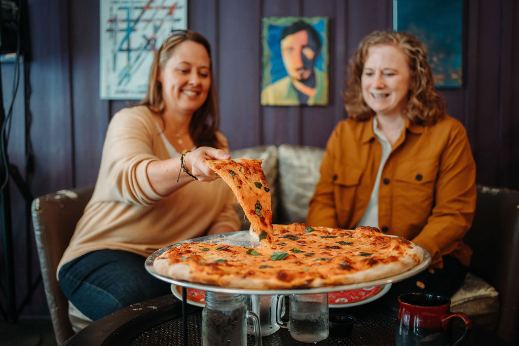 Two smiling women sit at a table with a large pizza. One woman reaches to take a slice while the other looks on. They are seated indoors with artwork visible in the background. Two beverages are also on the table.