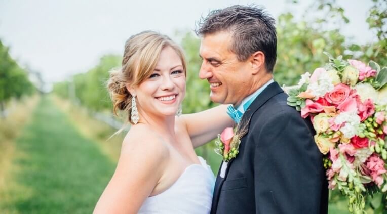 A bride in a white dress and a groom in a black suit with a turquoise bow tie stand outdoors. The bride, smiling happily at the camera, holds a bouquet of pink and white flowers. The groom, looking at the bride, shares a joyful moment with a greenery backdrop.