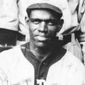 A historical black-and-white photograph of a man wearing a vintage baseball uniform, including a cap and a jersey. The man is looking directly at the camera with a neutral expression. The background shows parts of other individuals in similar attire.