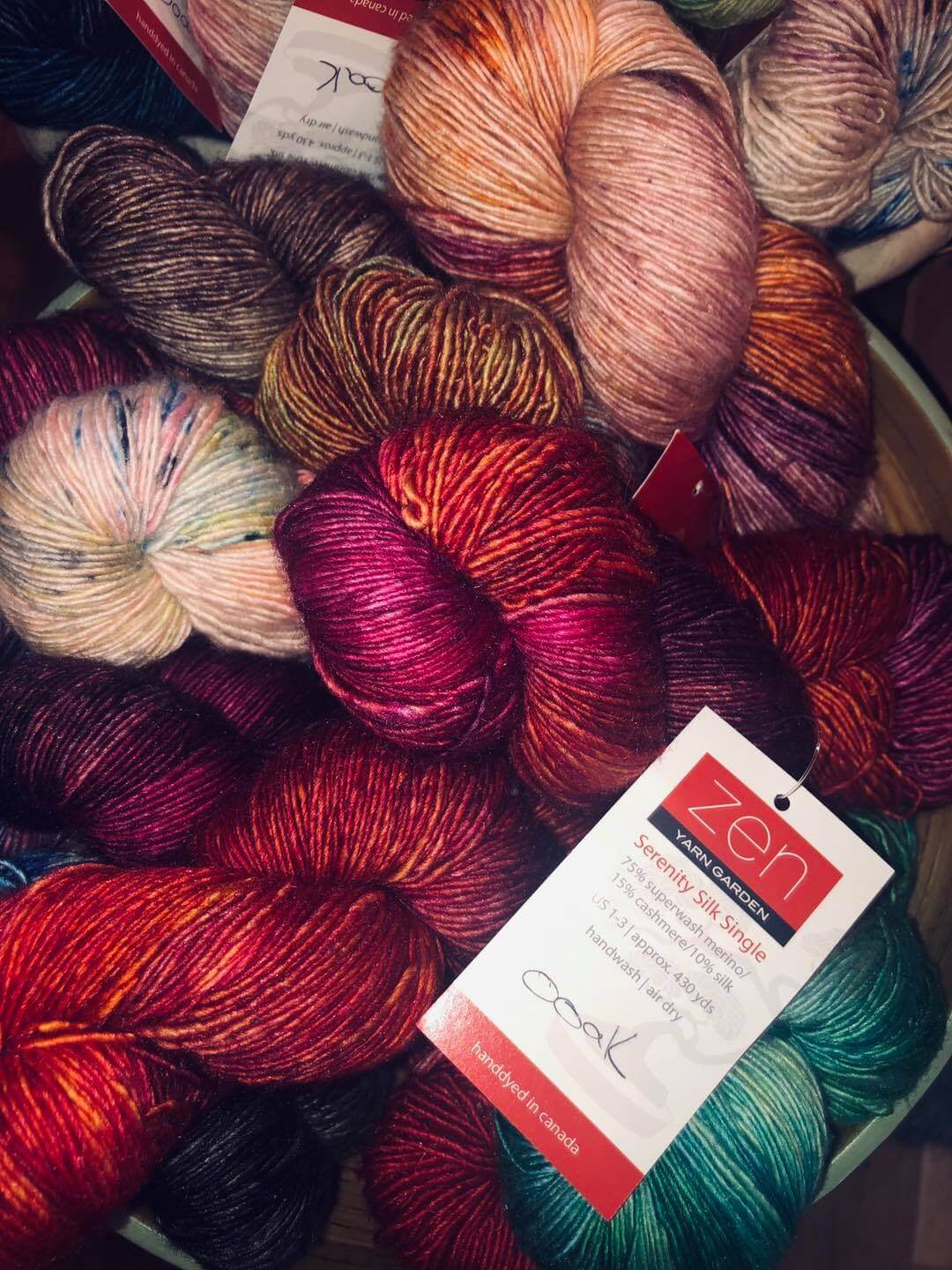 A pile of colorful skeins of yarn of various shades including red, pink, green, brown, and multi-colored ones. Each skein has a label reading 