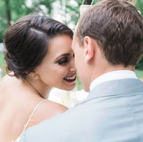 A couple sharing an intimate moment, with their faces close together and smiling. The woman has dark hair in an updo and is wearing a white dress, while the man has short, light hair and is dressed in a grey suit. They appear to be outdoors with greenery in the background.