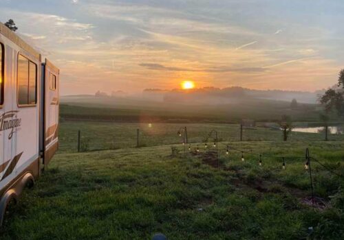 A scenic sunrise view over a foggy field with a camper trailer parked on a grassy area to the left. The sky is painted with hues of orange and pink as the sun rises, and string lights are draped along a fence in the foreground. Trees and a pond are in the distance. Image