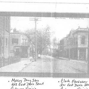 Black and white photo of a quiet tree-lined street with storefronts on either side. Visible signs include 