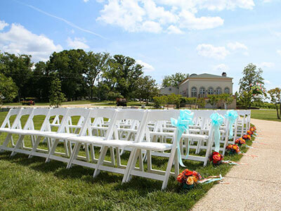 Rows of white folding chairs set up outdoors on a grassy lawn, decorated with turquoise bows and orange flowers, ready for a wedding ceremony. In the background, there are trees and a building under a sunny, blue sky with scattered clouds.