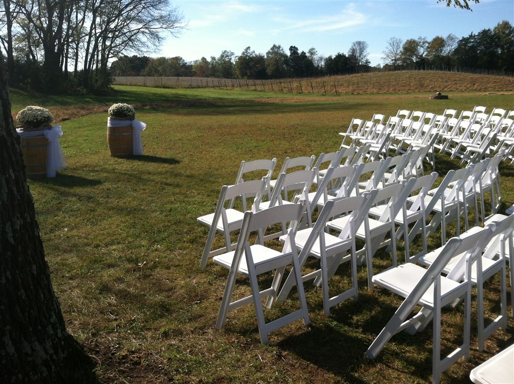 An outdoor wedding setup with rows of white chairs arranged in a grassy field. Two large flower arrangements sit atop wooden barrels at the front, marking the aisle. Trees and a vineyard are visible in the background under a clear, blue sky.