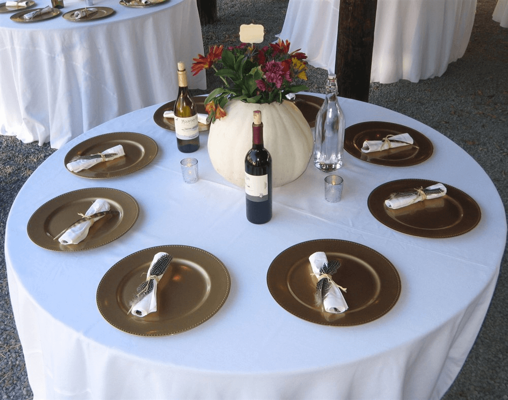 A round table set for six, covered with a white tablecloth. Each place has a gold plate and a white napkin. The centerpiece is a white pumpkin filled with colorful flowers. Three bottles, including wine and water, are placed on the table, along with small candles.