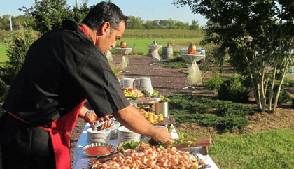 A chef in a black shirt and red apron arranges food on a long, outdoor buffet table. The table is set with various dishes, including a large platter of shrimp. The outdoor setting features greenery, trees, and a clear sky in the background.