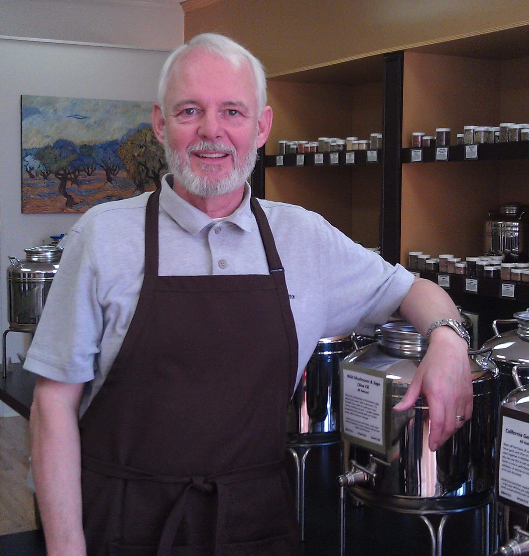 A bearded man wearing a brown apron over a gray polo shirt stands in front of stainless steel containers in a store. He is smiling and resting one arm on a container. Shelves behind him are stocked with small jars. A painting hangs on the wall in the background.