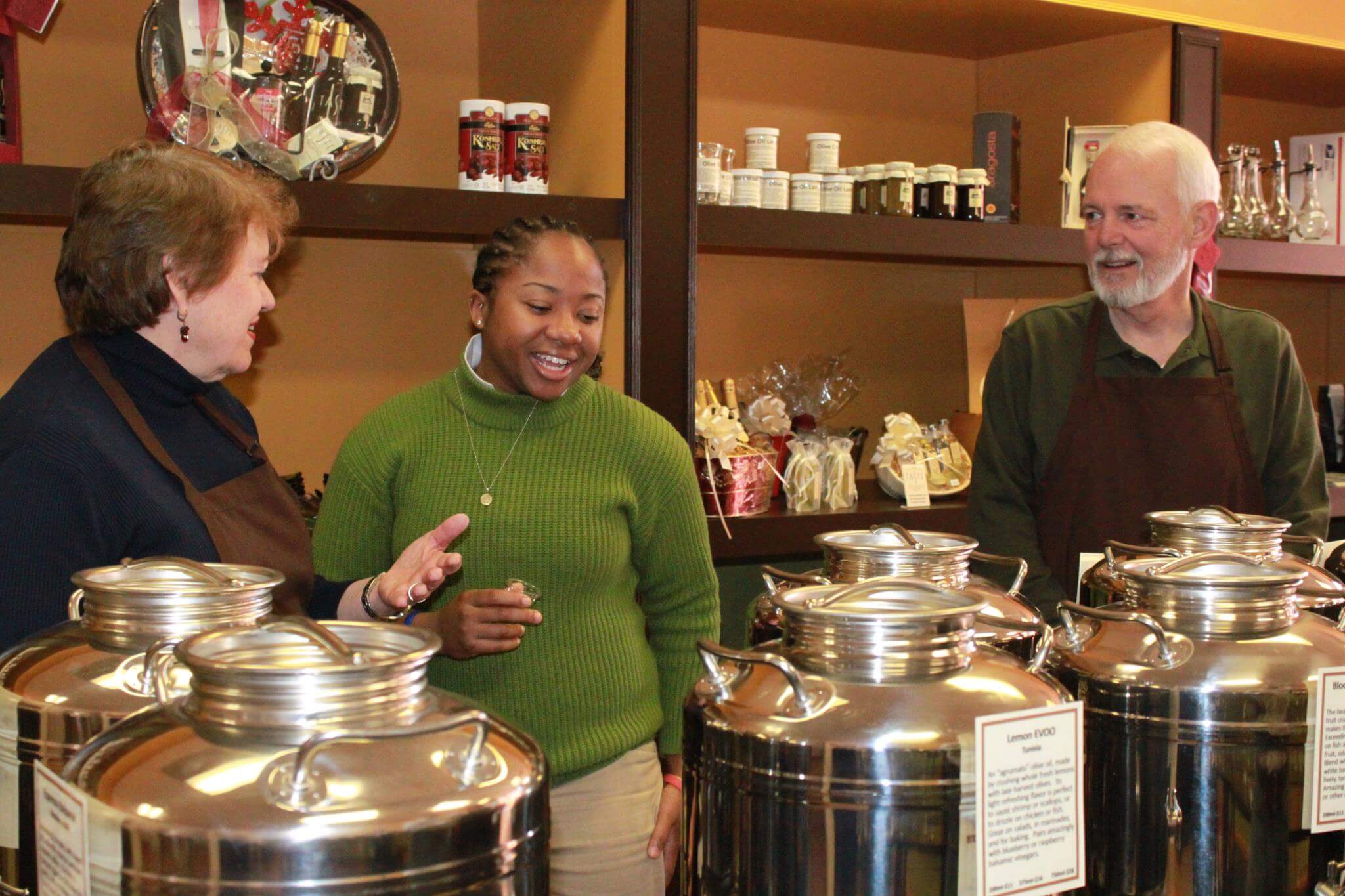 Three people wearing aprons stand behind a counter with stainless steel containers. The woman on the left is gesturing towards the woman in the middle, who is smiling. The man on the right is also smiling. Shelves with various goods are visible in the background.