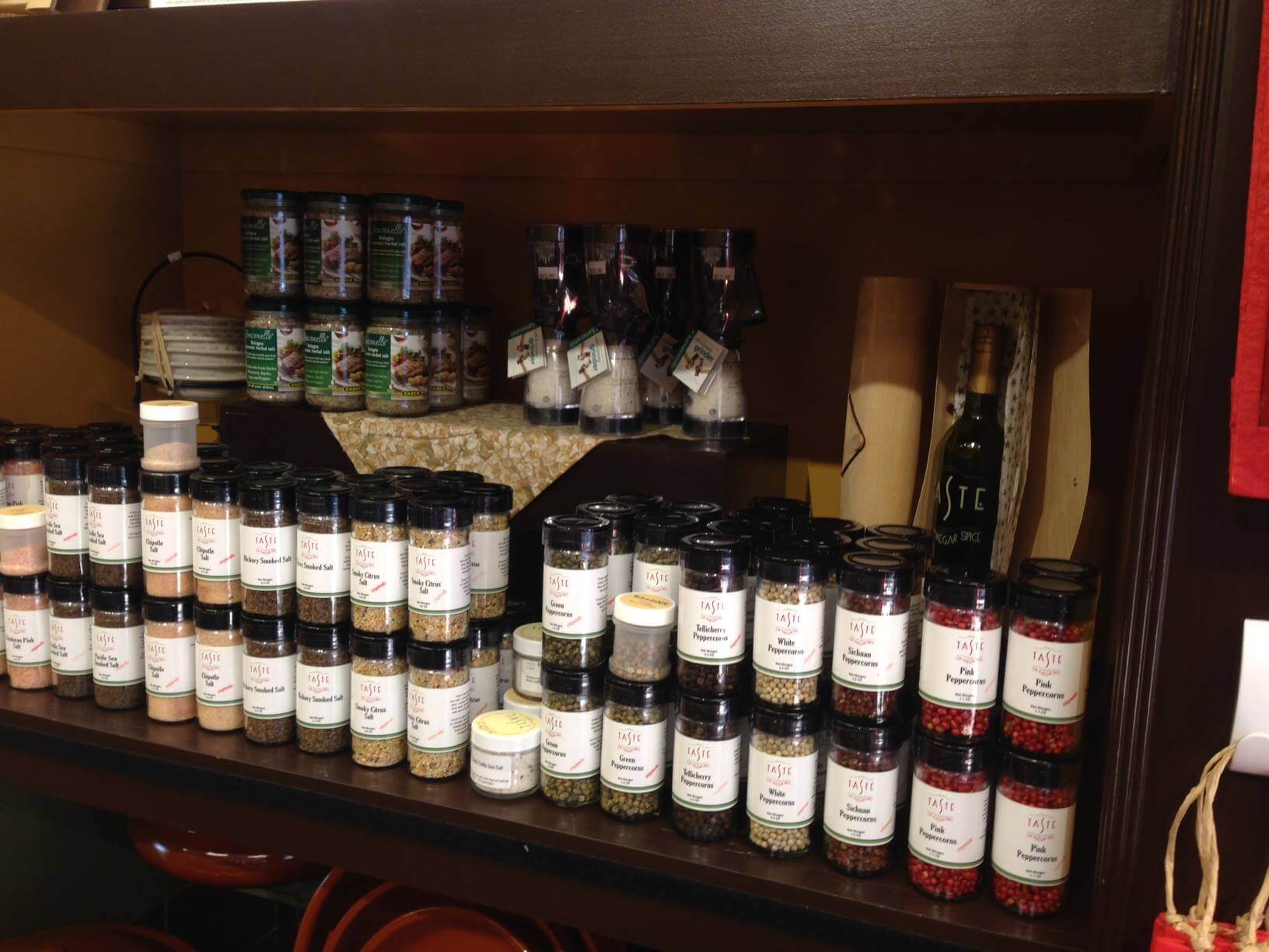 A shelf in a store displaying various jars of spices and seasonings. There are neatly arranged labeled jars, some containing peppercorns and salts. Above them are more spice containers and some packaged items, including a bottle of olive oil on the right.