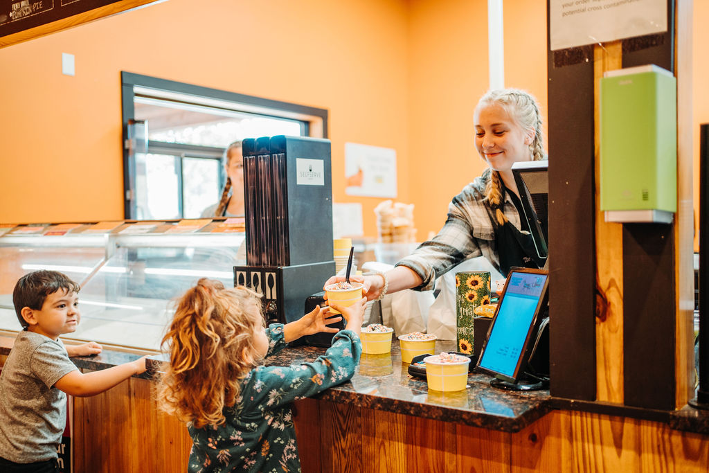 A young woman serves a cup of frozen yogurt to a child with red hair at a yogurt shop. Another child with brown hair stands beside the first, looking at the counter. The setting has bright orange walls, a wooden counter, and a yogurt machine.
