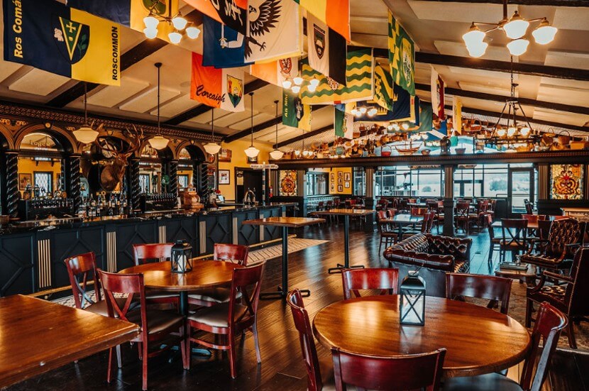 A warmly lit pub with wooden tables and chairs, and a long bar adorned with various flags hanging from the ceiling. Lantern-style lights provide a cozy atmosphere. The space has a mix of seating options, including tables and cushioned seats with a view towards the entrance.