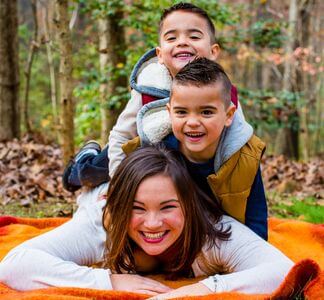 A woman lies on an orange blanket, smiling, with two young boys playfully stacked on her back. They are outdoors in a wooded area with fall leaves scattered around, enjoying a fun moment together.
