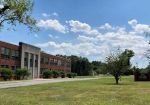 A two-story brick building with large windows, surrounded by green grass and trees, under a partly cloudy blue sky. A sign on the right side reads 
