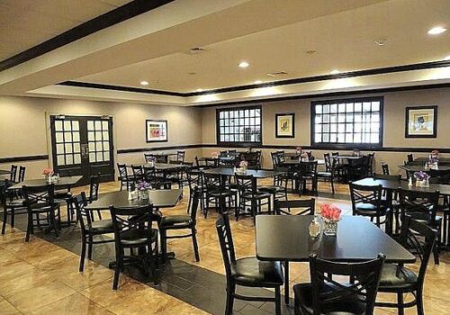 A spacious, well-lit restaurant interior with multiple empty black tables and chairs arranged neatly. Framed pictures adorn the beige walls, and there are windows with black frames. Small vases with flowers are placed on each table. Image