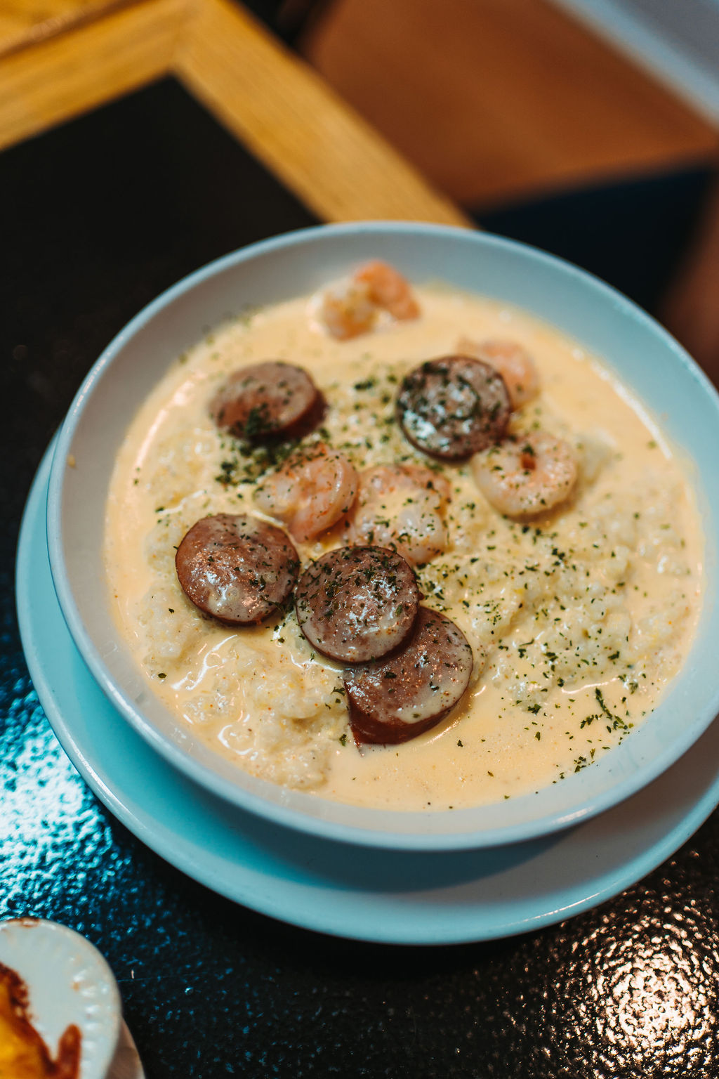 A white bowl on a dark surface filled with creamy grits, topped with shrimp and slices of sausage, seasoned with herbs and spices.