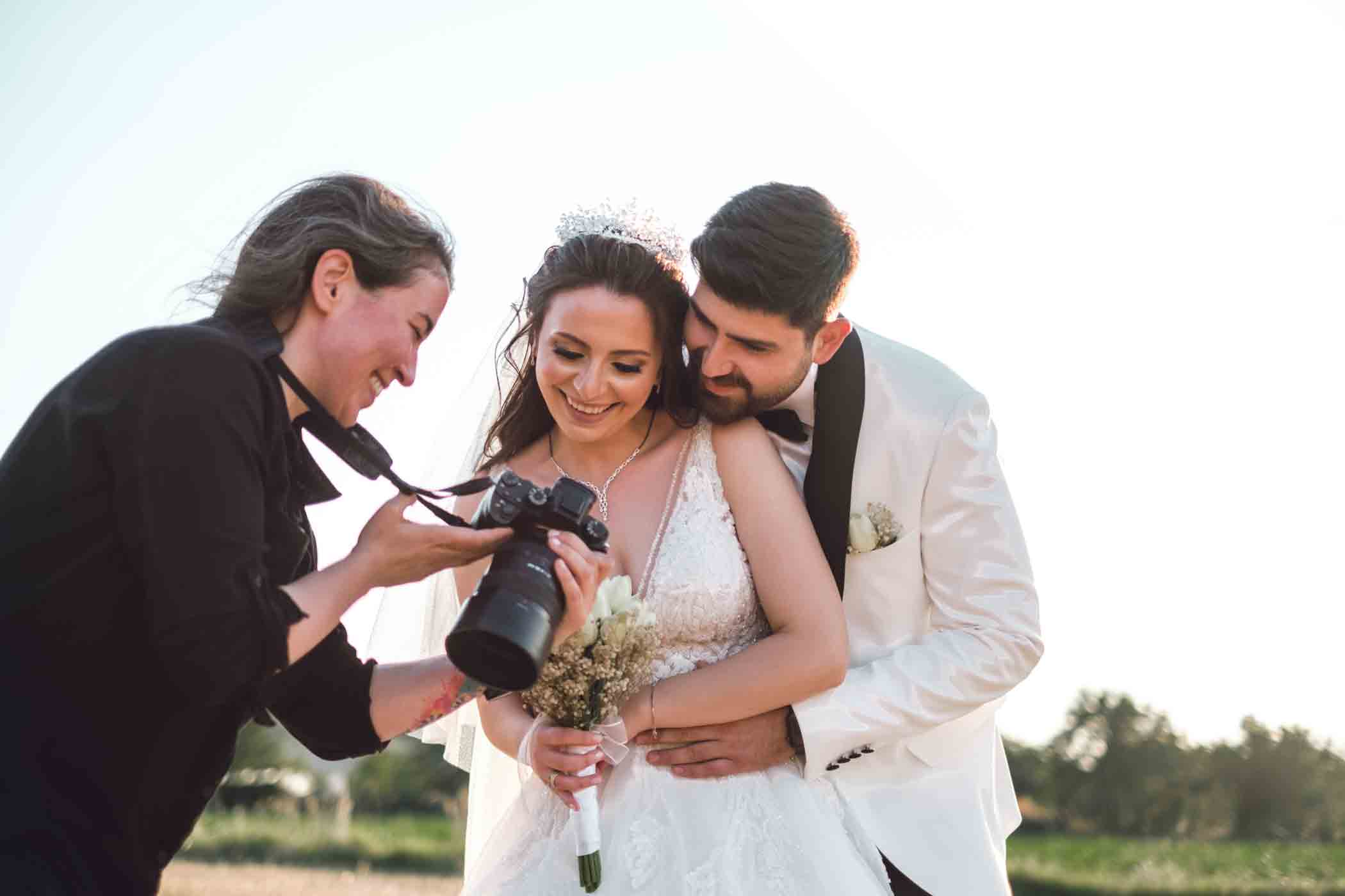 A newlywed couple in wedding attire embraces and smiles while looking at photos on a camera with a photographer dressed in black. The bride holds a bouquet and wears a tiara. They are outdoors in a natural setting under a clear sky.