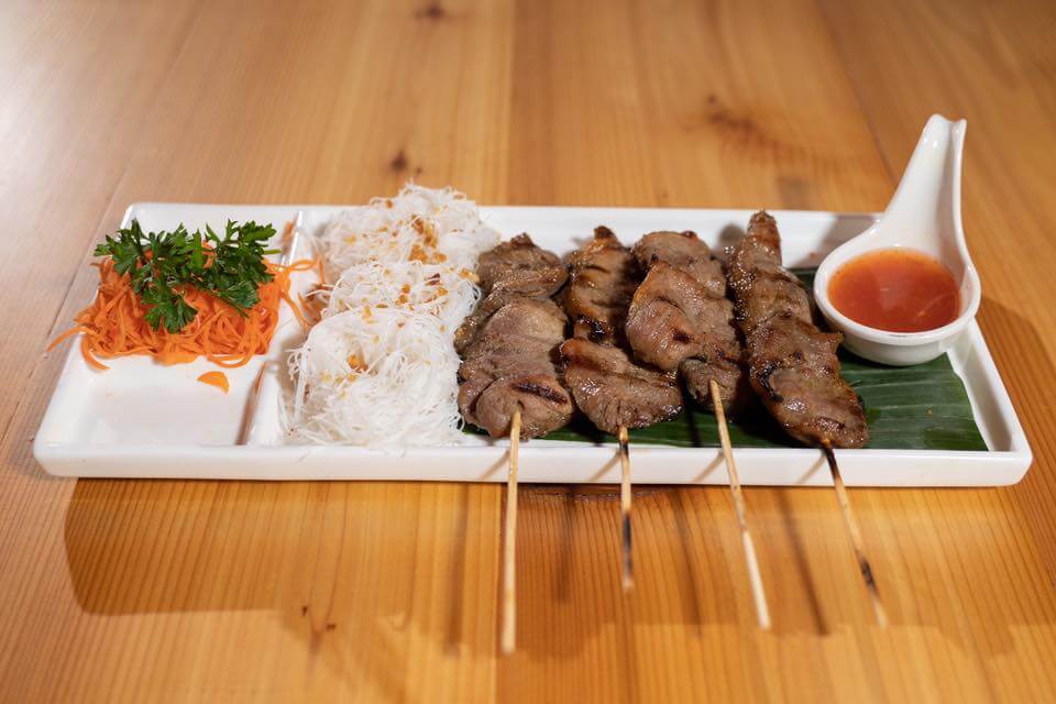 A rectangular white platter with three sections holds grilled meat skewers, a small mound of shredded carrots and greens, and thin rice noodles topped with fried onions. A small white sauce dish containing a red dipping sauce is set on the platter.