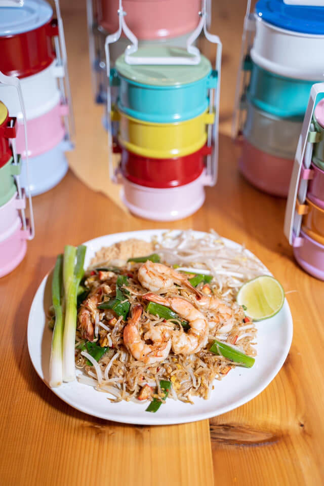 A plate of shrimp noodles garnished with greens and a lime wedge is set on a wooden table. Behind the plate, there are several colorful stacked tiffin lunchboxes. The overall ambiance suggests an appetizing meal ready to be enjoyed.