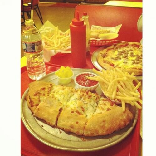 A table with various types of food: a plate with a sliced calzone, a small cup of marinara sauce, pepperoncini, and a side of fries; a bottle of water; a squeeze bottle of ketchup; a basket of fries and chicken tenders; and a small pizza in the background.
