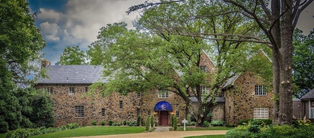A large stone building with a pitched roof is partially obscured by tall trees. The entrance features a purple awning with white text. A gravel path leads to the entrance, and the building is set against a backdrop of green foliage and a cloudy sky.