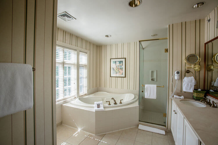 A spacious bathroom with beige vertical striped wallpaper features a corner jacuzzi tub near a window with blinds. There's a glass-enclosed shower to the right of the tub, and a countertop with a sink, vanity mirror, and towel rack on the opposite wall.