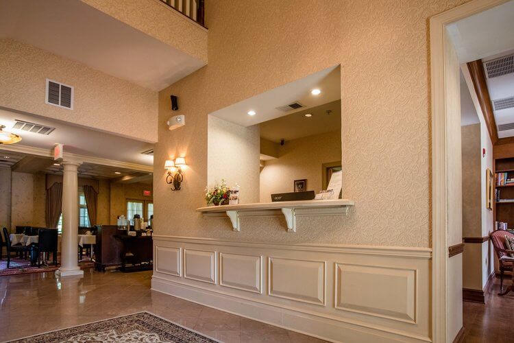 A warmly lit hotel lobby with beige textured walls and an elegant front desk. The area features a small check-in window with a floral arrangement and brochures. On the left, tables and chairs are visible near a column, and a patterned rug adorns the tiled floor.
