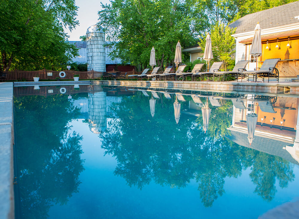 A serene swimming pool with clear blue water reflects the surrounding trees and sky. Lounge chairs with umbrellas line one side of the pool, set against a backdrop of greenery and a white building. The scene is calm and inviting, suggesting a peaceful retreat.