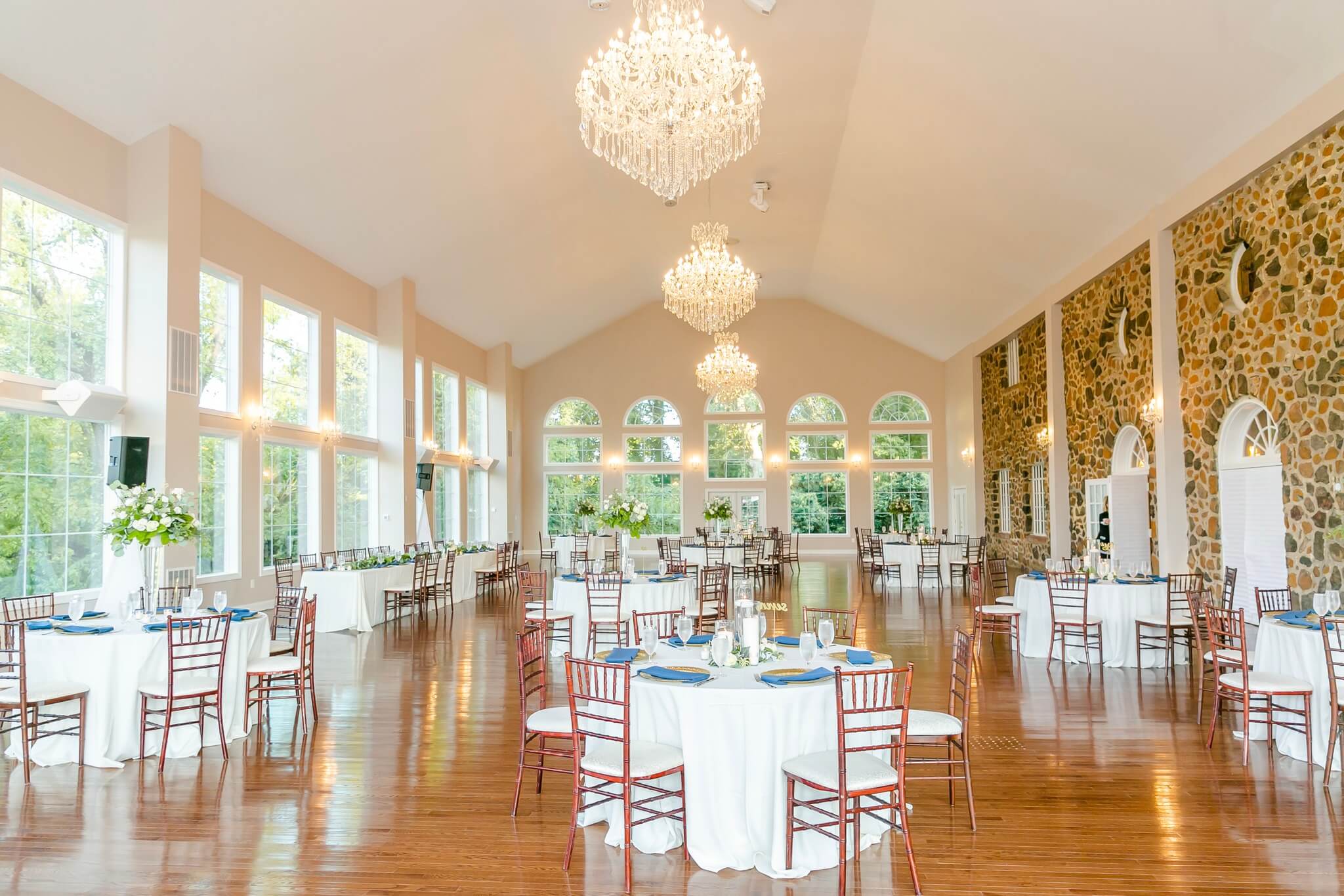 A spacious, elegant event hall with high ceilings and large windows. The room features chandeliers, wooden floors, stone walls on one side, and round tables set with white tablecloths and wooden chairs. The tables have floral centerpieces and table settings.