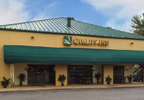 A front view of Quality Inn hotel featuring a green roof and a large green awning extending over the entrance. The building is painted in neutral tones, and there are small potted plants flanking the door. A small outdoor staircase is visible on the right side. Image
