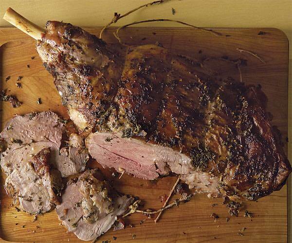 A roasted leg of lamb on a wooden cutting board, seasoned with herbs and spices. Portions of the lamb have been sliced, revealing the tender, juicy inside. The image highlights the textural contrast between the crusty exterior and succulent meat.