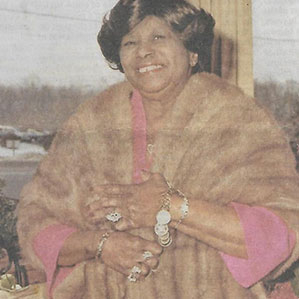 An older woman smiles warmly while wearing a light brown fur coat over a pink outfit. She is standing in front of a window with a snowy landscape outside and has multiple rings and bracelets on her hands, one arm crossed over her chest.