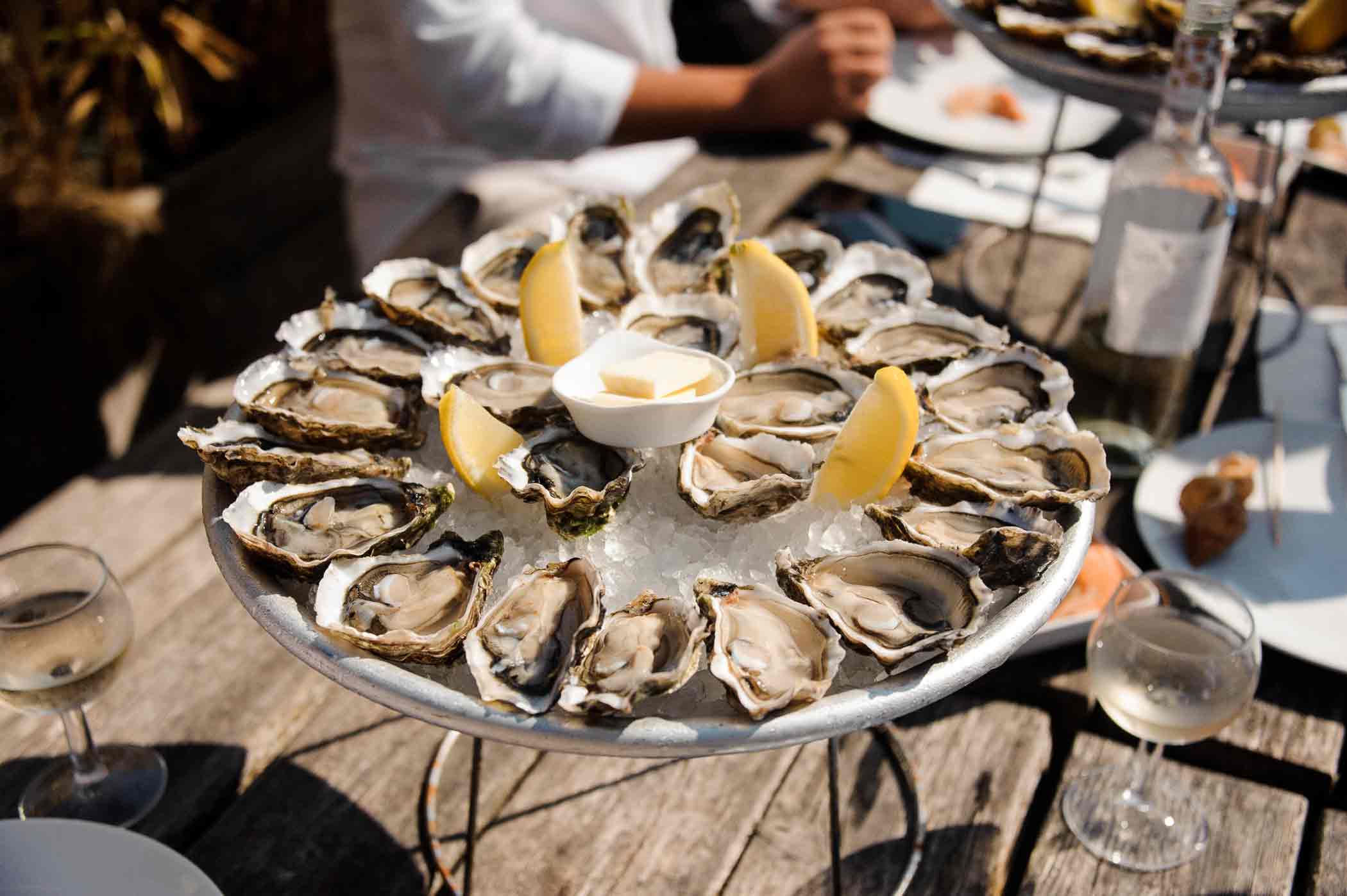A round platter filled with ice and arranged with opened oysters, lemon wedges, and a central bowl of dipping sauce is placed on a wooden table. Two glasses of white wine and plates with food are also present. The hands and part of a person are visible in the background.