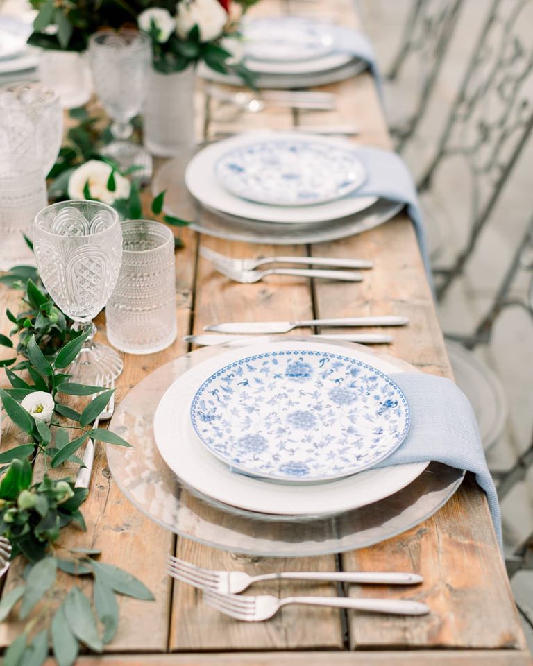 A rustic wooden table is beautifully set with white plates adorned with blue floral patterns. Clear crystal glassware and polished silverware are neatly arranged. Light blue napkins and a green leafy garland complete the elegant, nature-inspired tablescape.