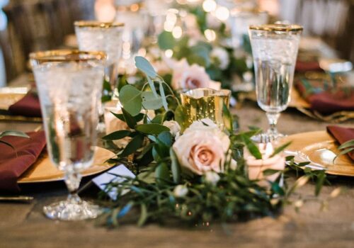 A close-up of an elegantly set dining table decorated with pink roses, green leaves, and candles. The table features crystal glasses filled with water, gold-rimmed plates, and dark red napkins, creating a sophisticated and romantic atmosphere. Image