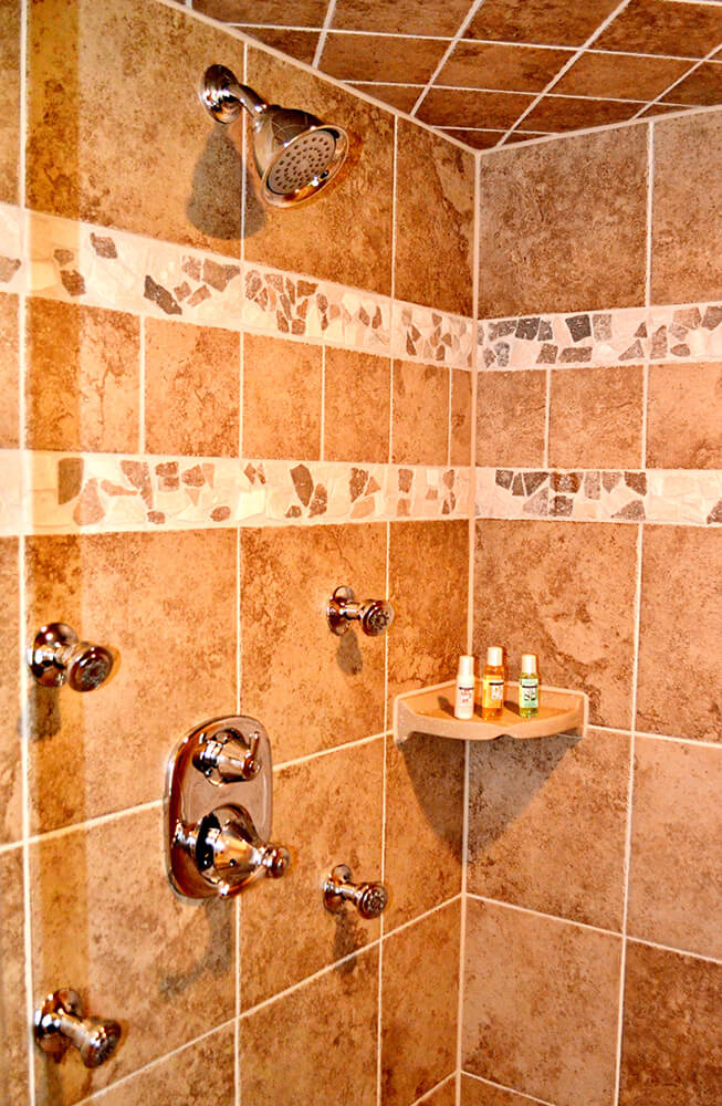 A tiled shower with brown and beige mosaic patterns on the walls and ceiling. It features multiple chrome-finished showerheads and knobs. On a small corner shelf, there are three small bottles of toiletries.