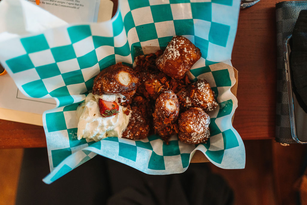 A basket lined with blue and white checkered paper holds powdered sugar-covered fried pastries, served with a dollop of cream and sprinkled crumbs. The basket is placed on a wooden table with part of a book and a black item visible beside it.