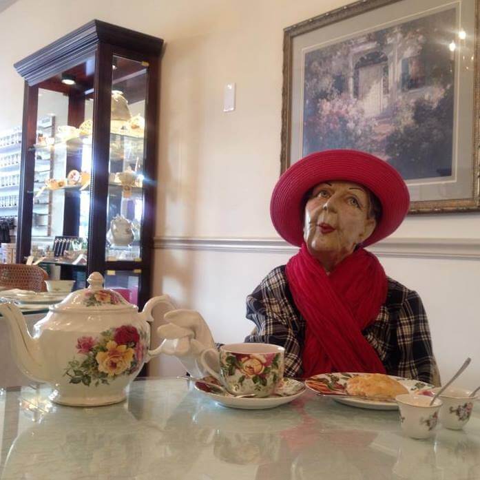 A mannequin dressed in a plaid outfit with a red scarf and red hat is seated at a table set for tea. The table features a floral teapot, teacup, and a plate with a pastry. Behind, there's a glass display cabinet with various teapots and teacups, and a framed painting on the wall.