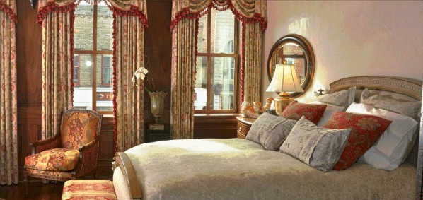 This image shows a luxurious bedroom with a large bed adorned with decorative pillows and an ornate bedspread. The room features patterned curtains, intricate wooden furniture, a vintage armchair, and a warm table lamp, creating an elegant and cozy atmosphere.