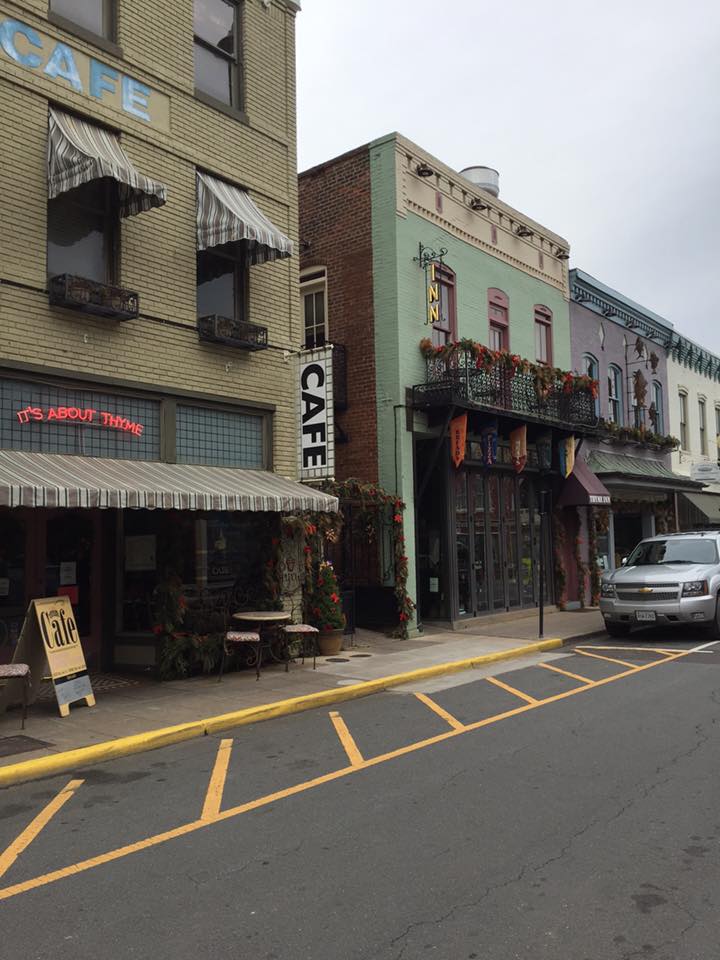 A quaint street scene featuring a row of colorful two-story buildings with awnings. Prominent signs include 