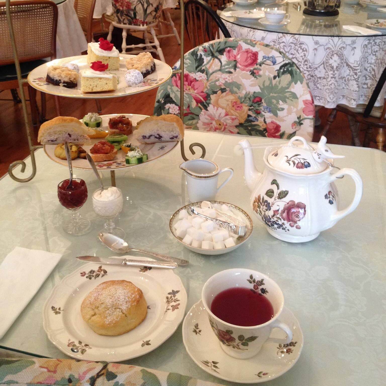 A table set for afternoon tea with floral china, a teapot, a teacup with red tea, a scone dusted with powdered sugar, and a tiered tray of assorted pastries and sandwiches. The table is covered with a pastel tablecloth and the chairs have floral cushions.