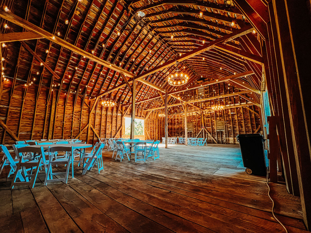 A rustic wooden barn with a high, arched ceiling is set up for an event. The interior is decorated with string lights and circular chandeliers. Several round tables and chairs are arranged throughout the space, predominantly in light blue.