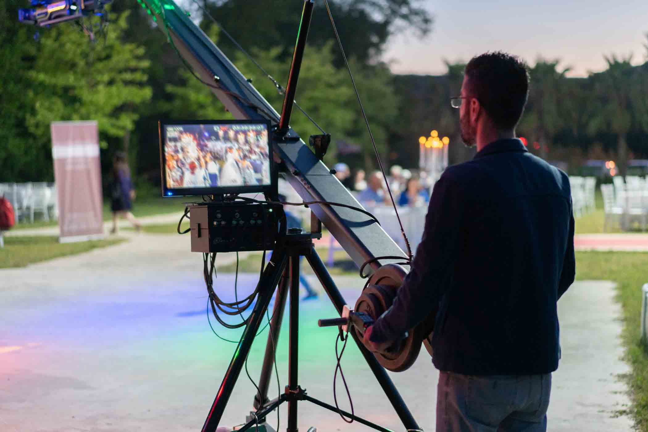 A person operates a camera crane with a monitor attached, capturing a live event outdoors. The scene is set during twilight with colorful lights illuminating the area. Empty chairs and blurred people are visible in the background.