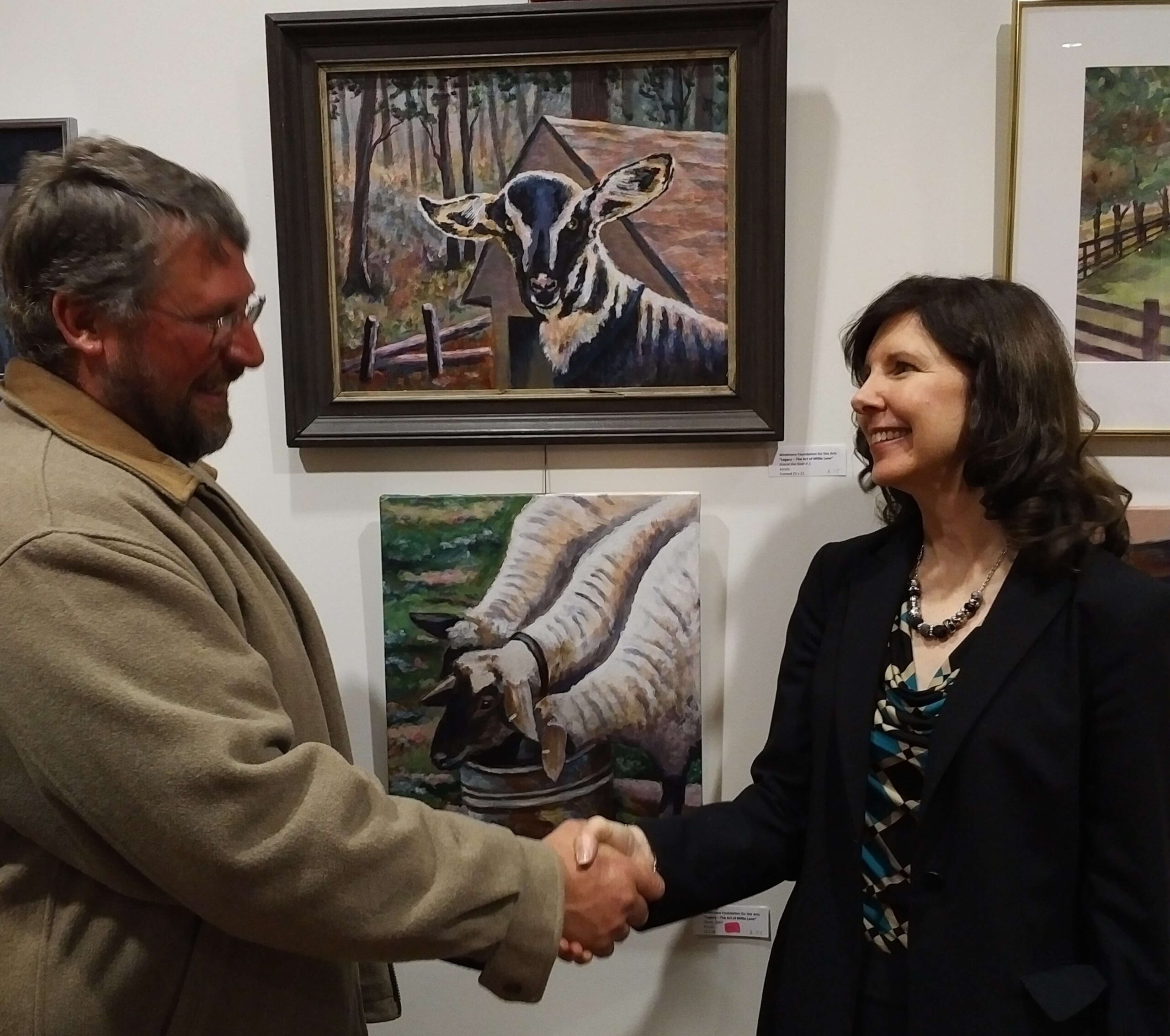 Two individuals shake hands in front of a wall displaying paintings, including a goat and two sheep. The person on the left wears glasses and a beige coat, while the person on the right has long dark hair and wears a black blazer over a patterned blouse. Both are smiling.